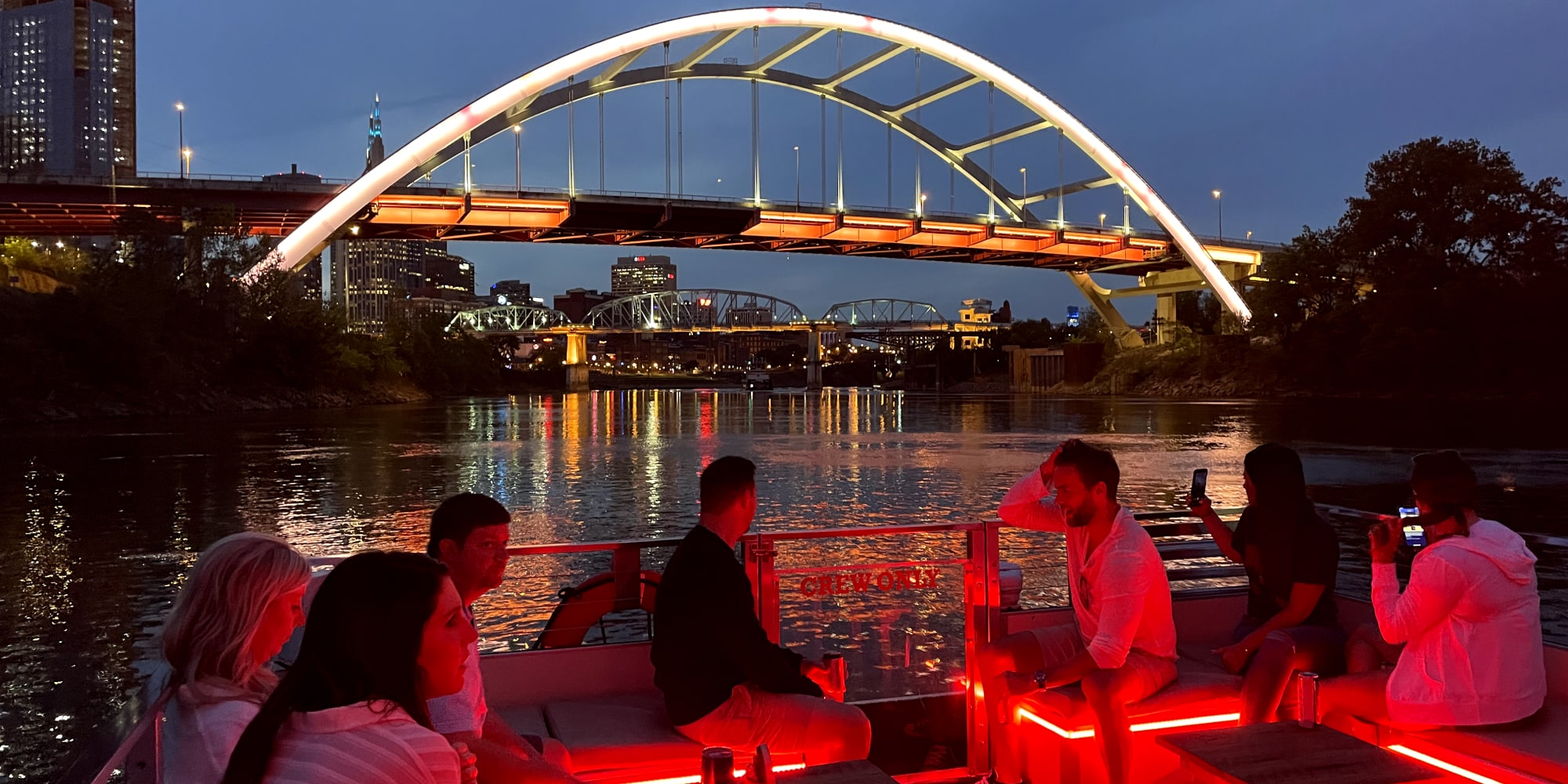 Group of men and women on a Nashville Water Taxi pontoon boat at night, with the Korean Veterans Memorial Bridge and Nashville skyline illuminated in the background. The boat’s red lights create a vibrant ambiance.