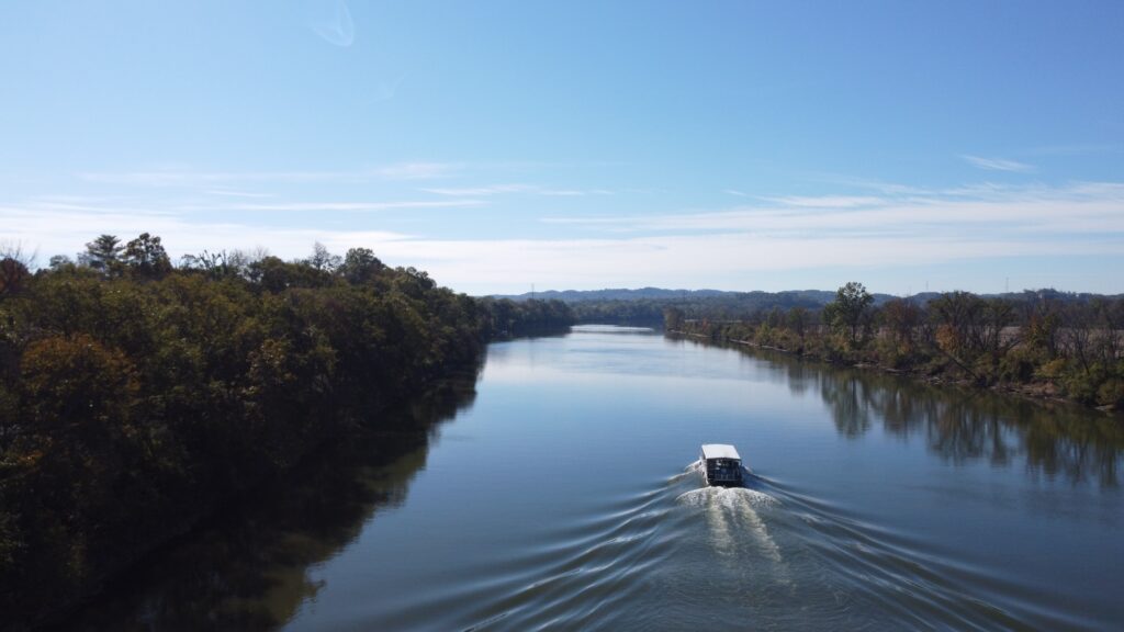 Nashville Water Taxi pontoon boat cruising the glassy Cumberland River, surrounded by trees and mountains under a blue sky.